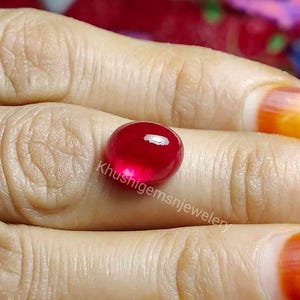 May include: A deep red gemstone with a smooth, polished surface. The gemstone is round and has a slight oval shape. It is held in a hand, and the background is blurred.