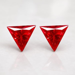 May include: Two vibrant red gemstones cut into a triangular shape. Each stone features a faceted surface that reflects light, creating a brilliant appearance. The stones are positioned side by side, showcasing their sharp, geometric edges and deep red color.