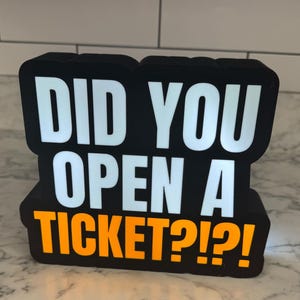 Puede incluir: Letrero LED en blanco y negro con el texto "DID YOU OPEN A TICKET?!?!" en letras blancas y naranjas. El letrero es rectangular y está sobre una encimera de mármol.