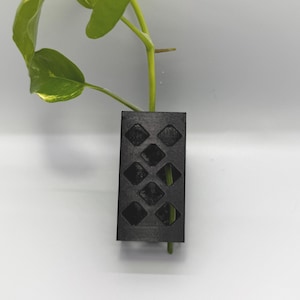 May include: A black, rectangular plant holder with a diamond pattern, holding a green plant with yellow-speckled leaves. The plant holder is made of a dark material and is designed to hold a single plant stem. The background is white.