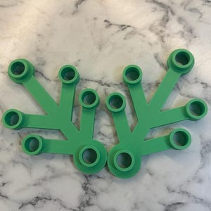 May include: Two green plastic components with a star-like design. Each component has a central circular base with five radiating arms, each ending in a circular opening. The objects are on a white and gray marble surface.