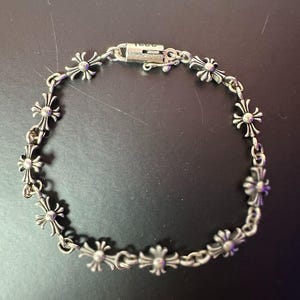 Può includere: Bracciale in argento con un motivo ripetuto di motivi floreali a forma di croce. Il bracciale ha una chiusura sicura ed è probabilmente realizzato in lega metallica. Il design è intricato e dettagliato, con un'estetica vintage o gotica.