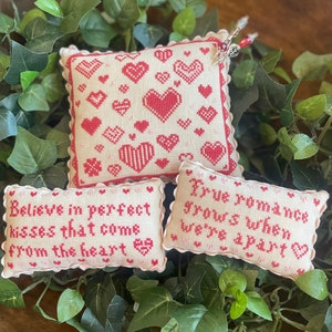 May include: Three small, white, embroidered cushion covers with red hearts and text. The cushions have scalloped edges and are decorated with red and white hearts. The text on the cushions reads "Believe in perfect kisses that come from the heart" and "True romance grows when we're apart".