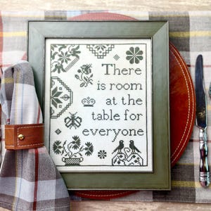 May include: A framed cross-stitch sampler with a green frame and a white background. The sampler features a floral design and the text "There is room at the table for everyone".
