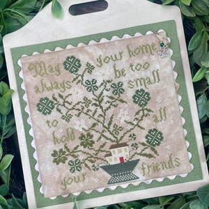 Puede incluir: Un sampler de punto de cruz con un esquema de color verde, blanco y marrón. El sampler presenta una planta en maceta con tréboles y el texto "Que tu hogar siempre sea demasiado pequeño para albergar a todos tus amigos".