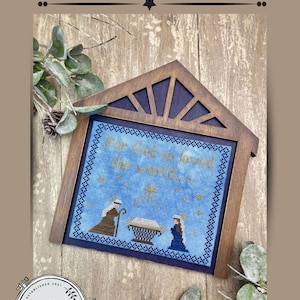 Może przedstawiać: Wzór haftu krzyżykowego zatytułowany "His Gift" przedstawiający scenę Bożego Narodzenia z tekstem "For God so loved the world". Projekt jest oprawiony w drewnianą ramę w kształcie domu, z nazwiskiem i logo artysty na dole.