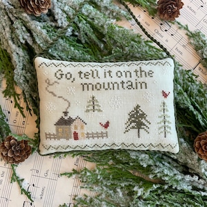 May include: A white pillow with green embroidery depicting a snowy winter scene with a small house, trees, and red birds. The text "Go, tell it on the mountain" is embroidered in green on the pillow.