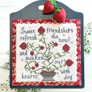 May include: A framed cross-stitch with the text "Sweet friendships refresh the soul and awaken our hearts with joy." The artwork features a floral design with red roses and white flowers. Two fabric strawberries are attached to the top.