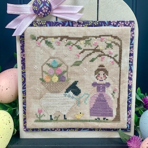 Könnte beinhalten: Eine Kreuzstich-Stickerei, die ein Mädchen in einem lila Kleid zeigt, das einen Korb mit bunten Eiern, ein weißes Schaf und ein gelbes Küken unter einem Baum mit rosa Blüten hält. Die Stickerei ist in einem Holzrahmen mit einer rosa Schleife und einem lila Knopf gerahmt.