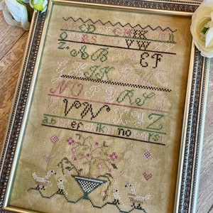 May include: A framed cross-stitch sampler with a green, brown, and pink color scheme. The sampler features the alphabet in a stylized font, along with a floral design and birds. The text reads "A B C D E F G H I J K L M N O P Q R S T U V W X Y Z".