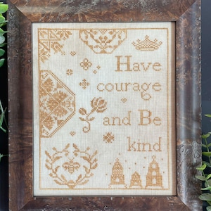 Op de afbeelding: Een ingelijst kruissteekkunstwerk met de woorden "Have courage and Be kind" in een decoratief lettertype. De kruissteek is in tinten bruin en beige, met bloemen- en geometrische patronen. De lijst is van donker, verweerd hout.