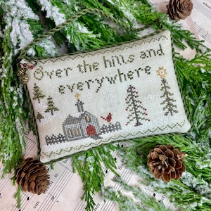May include: A white cross-stitch pillow with a winter scene and the text "Over the hills and everywhere". The pillow is surrounded by green pine branches and pine cones.
