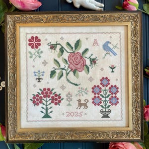 May include: A framed cross-stitch sampler with a floral design. The sampler includes a pink rose, a blue bird, and other flowers in red and blue. The year "2025" is stitched at the bottom. The frame is gold-coloured.
