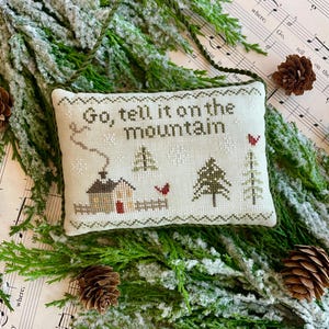 May include: A white cross-stitch pillow with a green and brown winter scene. The pillow reads "Go tell it on the mountain". The pillow is sitting on a sheet of music with a snowy evergreen branch.