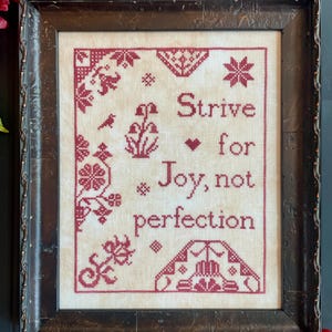 May include: Framed cross-stitch artwork with the words "Strive for Joy, not perfection" in burgundy thread. The design includes floral and geometric patterns in the corners and around the text. The frame is dark brown.