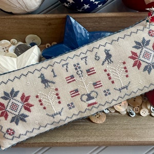 May include: A beige pillow with a cross-stitch design featuring red, white, and blue stars, flags, and the number 1776. The pillow is surrounded by wooden stars and buttons.