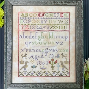 May include: A framed cross-stitch sampler with a vintage aesthetic. It features the alphabet, numbers, and the name "Frances Crayson" with the age "13" and the year "1863". The sampler is stitched in shades of red, green, blue, and purple.