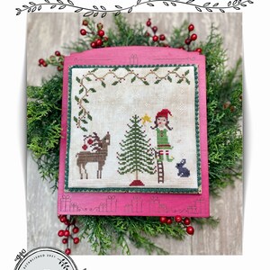 May include: Cross stitch pattern featuring a girl decorating a Christmas tree with a reindeer and a bunny nearby. The pattern is framed in a pink wooden frame with a white background and the text "Holly's Helpers" at the top.