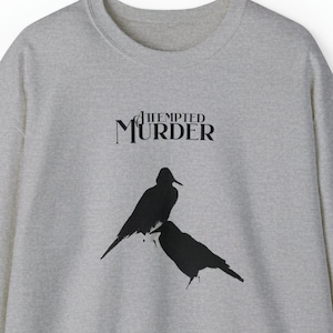 Peut inclure: Un sweat-shirt gris avec le texte "Attempted Murder" et une silhouette noire de deux oiseaux sur le devant.