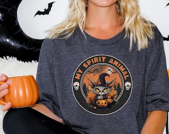 Puede incluir: Una camiseta gris con un gráfico negro y naranja de un gato con un sombrero de bruja y sosteniendo calabazas. El gráfico está rodeado por un círculo con el texto "My Spirit Animal".