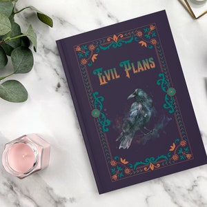 May include: A dark purple journal with a decorative border in shades of teal and orange. The journal features a black raven illustration and the text "Evil Plans" in orange lettering.
