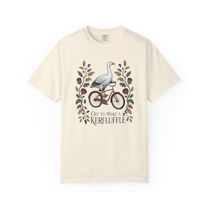 Könnte beinhalten: Cremefarbenes T-Shirt mit einer Gans auf einem Fahrrad. Die Gans ist grau und weiß, und das Fahrrad ist braun. Der Text "Off to make a Kerfuffle" ist in einer dekorativen Schriftart dargestellt, umgeben von floralen Elementen.