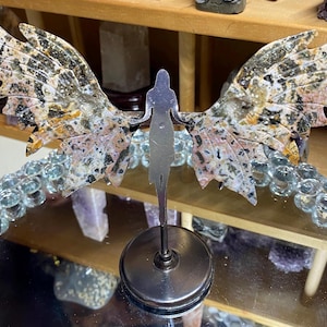 May include: A decorative angel wing sculpture with a silver silhouette figure. The wings are made of a speckled stone with shades of black, brown, and pink. The sculpture is mounted on a silver base and is surrounded by a strand of clear glass beads.