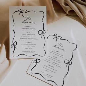 Op de afbeelding: Elegante trouwmenukaarten met een zwarte lintontwerp-rand. De kaarten tonen de tekst "The Menu" in cursief, samen met menu-items voor voorgerechten, hoofdgerechten en desserts. Gedrukt op wit papier.