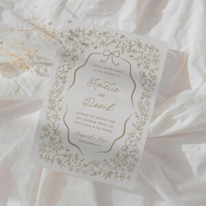 May include: A wedding invitation with a floral design in gold ink. The invitation includes the names "Amelia and David" and the wedding date and time. The text is framed by a decorative border of flowers and leaves.