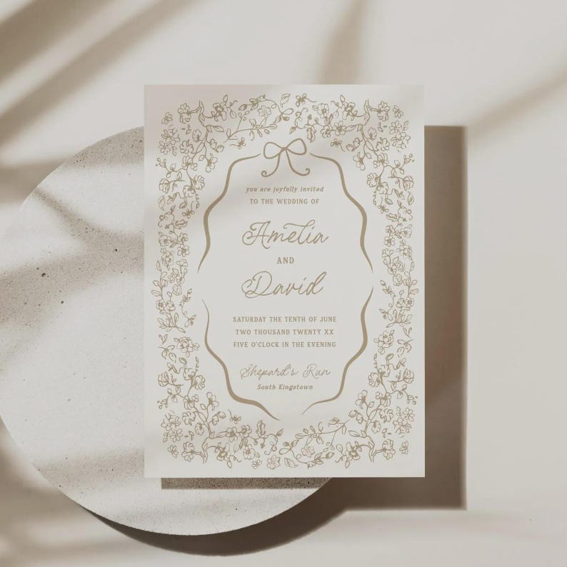 May include: A wedding invitation with a floral border and an oval frame. The invitation is cream-colored with gold-colored text that reads "Amelia and David" and details about the wedding date and location. The invitation is on a white surface.