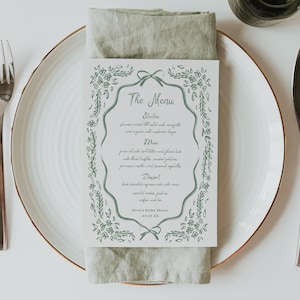 May include: A white plate with a gold rim, a fork, and a knife are set for a meal. A menu card with a green floral border and text is placed on a folded green linen napkin. The menu card reads "The Menu" and lists starters, main, and dessert.