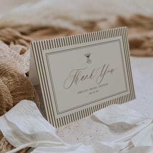 May include: A folded thank you card with a cream background and brown striped border. The card features the words "Thank You" in elegant script, along with the text "Amelia's Bridal Shower" and the date "08.15.XX". A small cocktail glass icon is at the top.