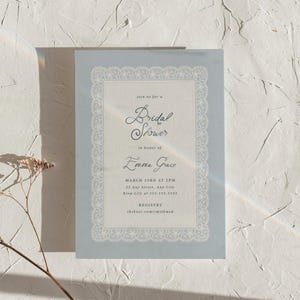 Può includere: Un invito per un bridal shower azzurro con un delicato bordo in pizzo. L'invito include il testo "join us for a Bridal Shower in onore di Emma Grace" e i dettagli dell'evento. Sono incluse anche le informazioni sulla registrazione.