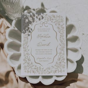 May include: A wedding invitation with gold floral accents and text. The invitation reads "Amelia and David" and includes the date and time of the wedding. The invitation is on a white background and is placed on a decorative white plate.