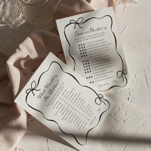 Pu&ograve; includere: Due fogli di carta bianchi con testo nero e un bordo decorativo a fiocco. Un foglio recita "What Would She Rather?" con un elenco di opzioni. L'altro recita "Over or Under?" con una serie di domande. Un bicchiere &egrave; sullo sfondo.