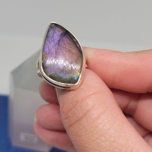 May include: A silver ring featuring a large, teardrop-shaped labradorite stone. The stone displays a shimmering play of color, with hues of purple, pink, and blue. The ring has a simple, elegant design, showcasing the natural beauty of the gemstone.