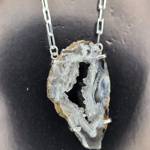 May include: A natural geode pendant necklace. The pendant is a slice of a geode, showcasing a mix of gray, white, and brown colors. The geode is suspended from a silver chain necklace.