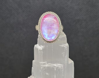 Pode incluir: Um anel de prata com uma pedra preciosa oval e iridescente. A pedra preciosa tem tons rosa, azul e branco. A banda do anel tem um design de corda torcida em torno da pedra preciosa. O anel é exibido em uma formação de cristal branca e em camadas contra um fundo escuro.