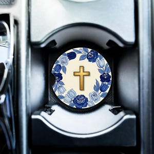 May include: A round car coaster featuring a gold cross in the center, surrounded by a wreath of blue flowers and leaves. The coaster is set in a car cup holder, with a neutral background.