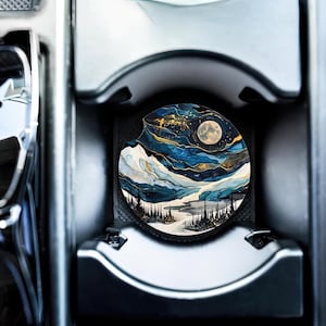 May include: A round car coaster with a blue, white, and gold abstract design featuring a full moon and a mountain range. The coaster is in a car cup holder.