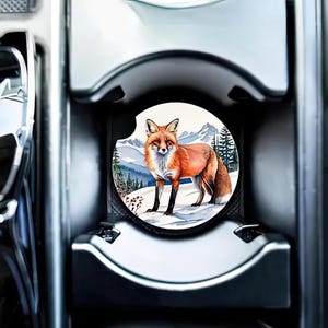May include: A round car coaster with a colorful illustration of a red fox standing in a snowy forest scene.