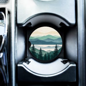 Può includere: Un sottobicchiere rotondo per auto con un paesaggio di montagne, alberi e un lago. Il sottobicchiere ha uno sfondo neutro con sfumature di verde e blu. Il sottobicchiere è inserito nel portabicchieri di un'auto.