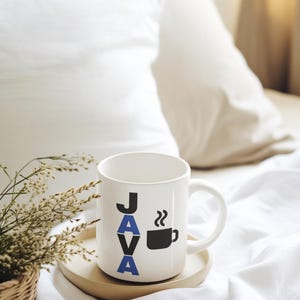 Pu&ograve; includere: Tazza da caff&egrave; in ceramica bianca con la scritta "JAVA" in lettere nere e blu e una grafica di tazza da caff&egrave; nera. La tazza &egrave; appoggiata su un vassoio di legno, con una piccola pianta in un cestino intrecciato a sinistra. Lo sfondo &egrave; un letto bianco.