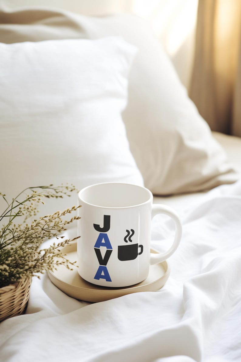 Pu&ograve; includere: Tazza da caff&egrave; in ceramica bianca con la scritta "JAVA" in lettere nere e blu e una grafica di tazza da caff&egrave; nera. La tazza &egrave; appoggiata su un vassoio di legno, con una piccola pianta in un cestino intrecciato a sinistra. Lo sfondo &egrave; un letto bianco.