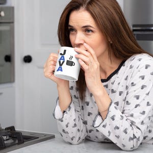 Pu&ograve; includere: Una tazza da caff&egrave; bianca con la scritta "JAVA" in lettere nere e blu e un disegno di tazza da caff&egrave;. La tazza &egrave; tenuta da una persona che indossa un top di pigiama bianco con un motivo a cuore nero. La persona sta bevendo dalla tazza.