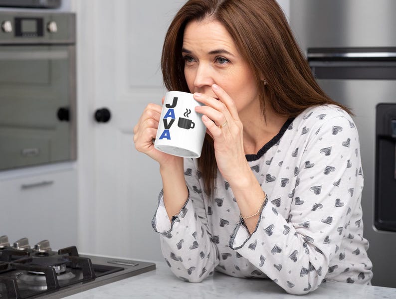 Pu&ograve; includere: Una tazza da caff&egrave; bianca con la scritta "JAVA" in lettere nere e blu e un disegno di tazza da caff&egrave;. La tazza &egrave; tenuta da una persona che indossa un top di pigiama bianco con un motivo a cuore nero. La persona sta bevendo dalla tazza.