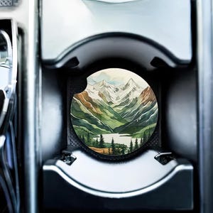 May include: A round car coaster with a watercolor painting of a mountain range with green trees and a lake. The mountains are snow-capped and the sky is a light blue.