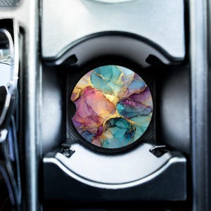 Peut inclure: Un dessous de verre rond pour voiture avec un motif abstrait dans les tons de turquoise, rose, violet et or. Le dessous de verre est placé dans le porte-gobelet d'une voiture, noir et argent.