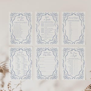Pu&ograve; includere: Sei biglietti per giochi per baby shower con bordi floreali blu e testo. Le carte includono giochi come "All Eyes on Mama" e "Over or Under". Le carte sono disposte su una superficie bianca con piante secche agli angoli.