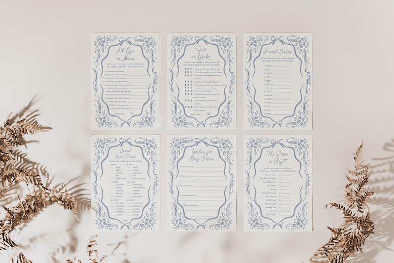 Pu&ograve; includere: Sei biglietti per giochi per baby shower con bordi floreali blu e testo. Le carte includono giochi come "All Eyes on Mama" e "Over or Under". Le carte sono disposte su una superficie bianca con piante secche agli angoli.